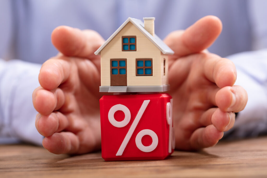 Close-up Of A Person's Hand Protecting House Model Over Red Cube With Percentage Symbol