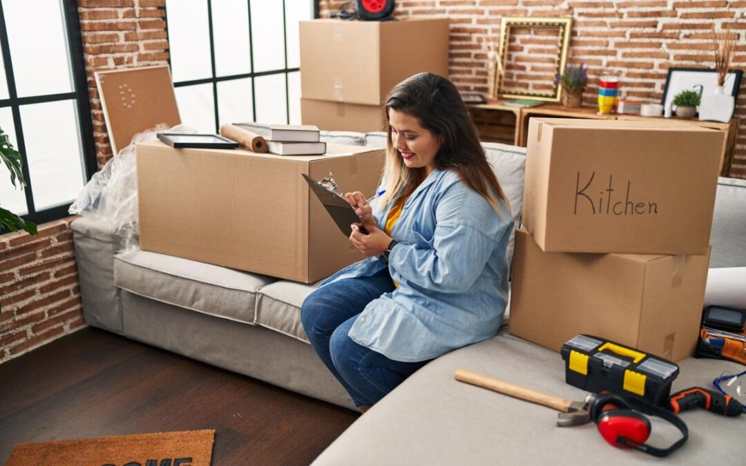 Woman checking off items on a clipboard as she sits in her new apartment, surrounded by boxes.