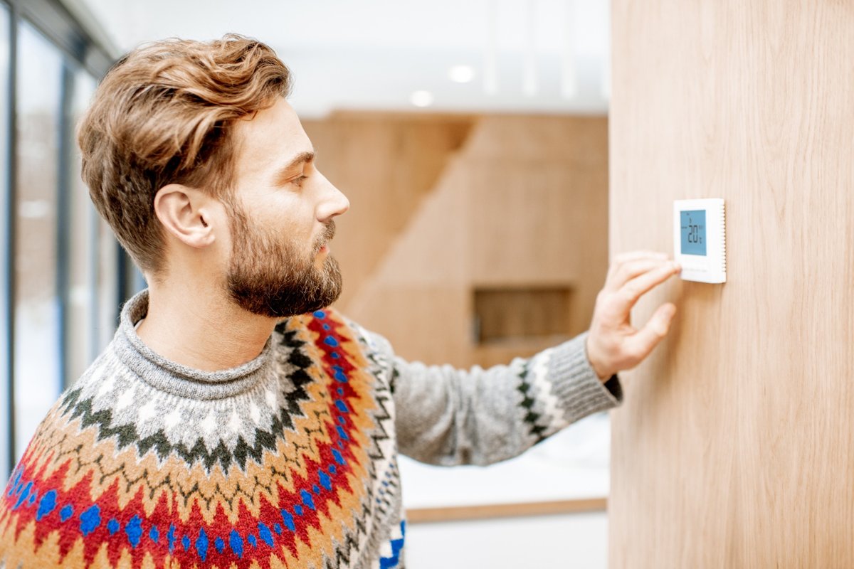 Man wearing a colorful sweater adjusting the thermostat that is on the wall in his living room.