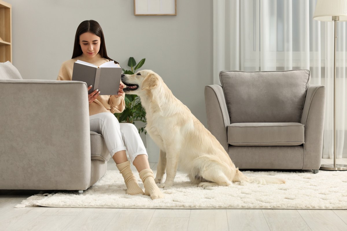 Female with fuzzy socks reading a book with her large dog sitting next to her on a cozy rug.