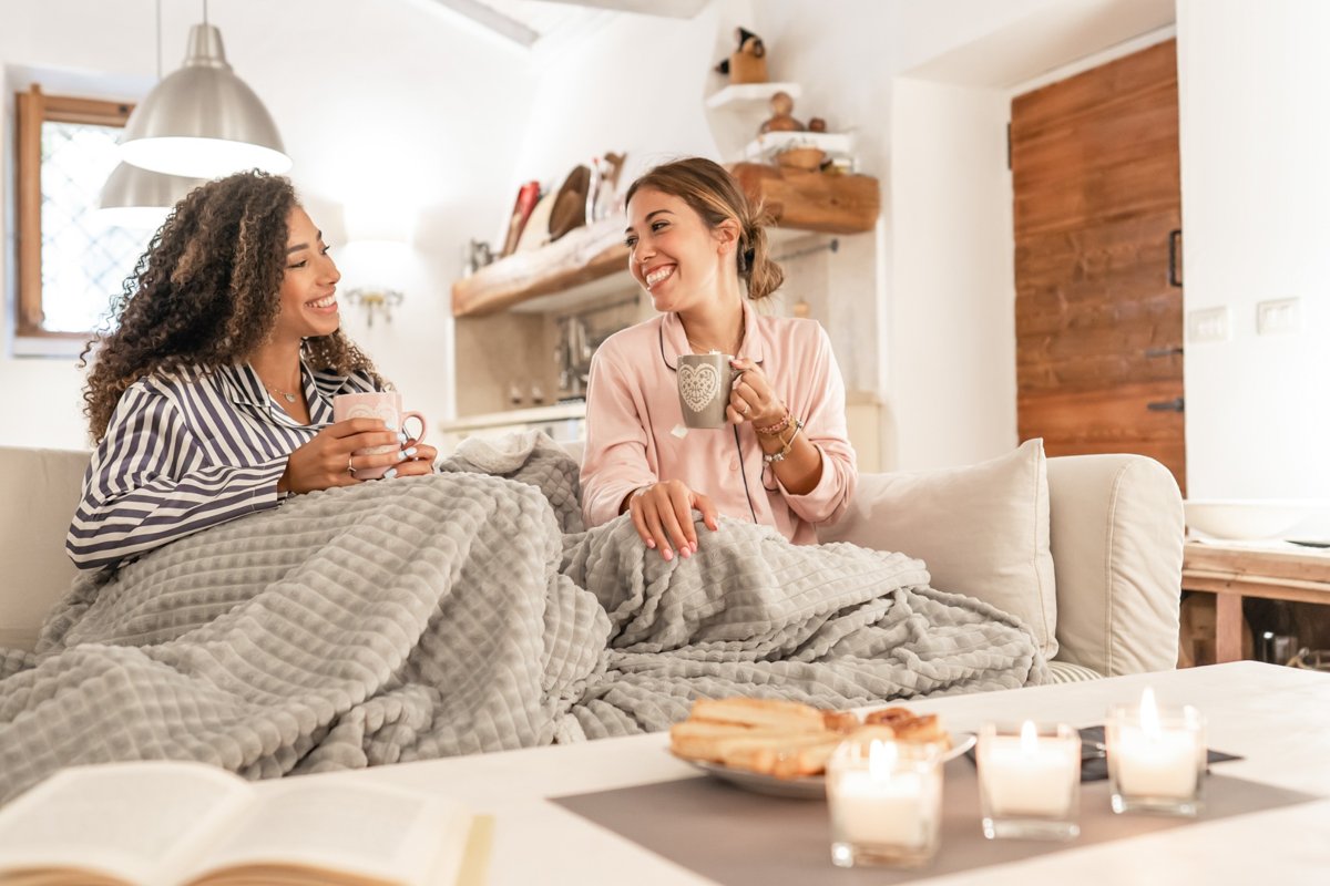 Two friends on a sofa with a blanket sharing hot tea and smiling.