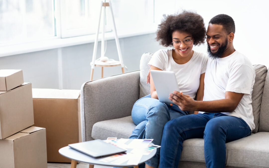 A young couple sits on a couch surrounded by moving boxes and looks at something on a tablet.